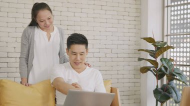 Mutlu Asyalı güzel aile çifti karı koca oturma odasındaki koltukta oturup gülerek evde dizüstü bilgisayarla çalışıyorlar. Kadın, iş sırasında adama kahve getirir..