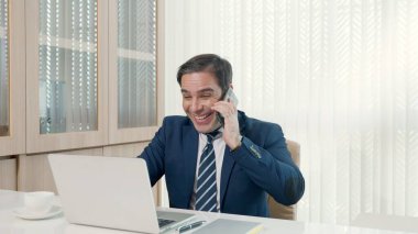 İşadamı ofisteki masa başı başarı kutlamalarında dizüstü bilgisayarını kullanarak oturuyor, gündelik ofiste mutlu bir iş adamı gibi gülümsüyor ve dizüstü bilgisayarla çalışırken telefonla konuşuyor.