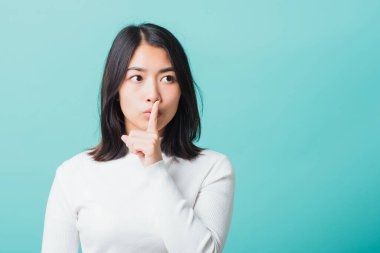 Dudaklarında işaret parmağı olan genç, güzel bir Asyalı kadın, sessiz bir kadın portresi, mavi arka planda izole edilmiş bir stüdyo çekimi, 