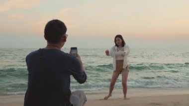 Mutlu bir adam kumsalda kadınlara fotoğraf çektirirken ve gün batımında tatilde sahilde birlikte eğlenirken, genç Asyalı kadın ve erkek yaz seyahatinde.