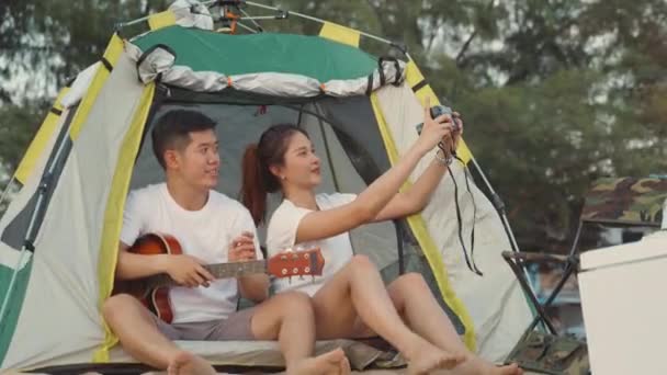キャンプ中の若い愛情のカップルはかなりカメラで写真を撮りながら 休日の休暇の時間にビーチのキャンプテントに座って日没 若いアジアのグループの女性と夏の旅行の屋外で男 — ストック動画