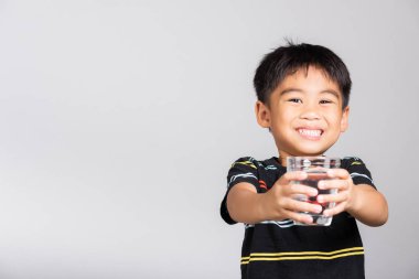 5-6 yaşlarında küçük tatlı bir çocuk beyaz arka planda izole edilmiş beyaz çekimde camdan tatlı su içerek gülümsüyor. Asyalı çocuklar anaokulu, günlük yaşam sağlığı.