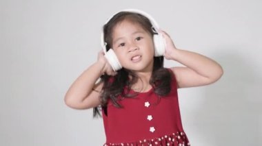 3-4 yaşlarında tatlı bir kız çocuğu. Beyaz arka planda izole edilmiş kablosuz kulaklıklarla müzik dinliyor. Mutlu Asyalı çocuklar gülerek müzik dinliyor.