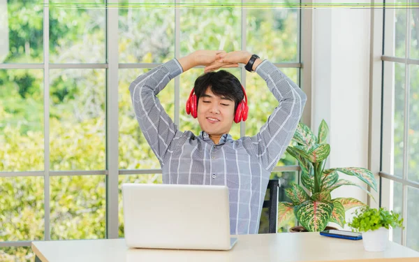 アジアの若いビジネスマンは 自宅のオフィスで赤いヘッドフォンで音楽を聞いて笑顔 幸せなビジネスマンは 机の上に座って リラックスした着用のワイヤレスヘッドフォンは 音楽を聴くとラップトップコンピュータで作業を楽しむ — ストック写真