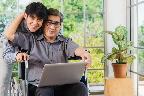 坐在轮椅上 带着手提电脑的亚洲资深残疾商人与办公室里的团队一起讨论 老父亲坐在轮椅上 儿子在笔记本电脑上进行视频电话会议 — 图库照片