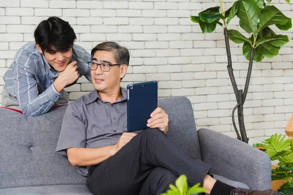 拥有数码平板电脑的亚洲资深商人与年轻的在位团队一起讨论 爸爸和儿子坐在沙发上聊天 在家里客厅里用平板电脑举行电话会议 — 图库照片
