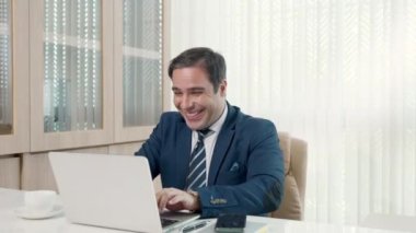 İşadamı ofisteki masa başı başarı kutlamalarında dizüstü bilgisayarını kullanarak oturuyor, gündelik ofiste mutlu bir iş adamı gibi gülümsüyor ve dizüstü bilgisayarla çalışırken telefonla konuşuyor.