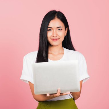 Mutlu Asyalı güzel bir genç kadının portresi dizüstü bilgisayarla gülümseyen yüzünü tutuyor, kameraya bakıyor, stüdyo görüntüsü pembe arkaplanda, kopyalama alanı ile izole edilmiş.