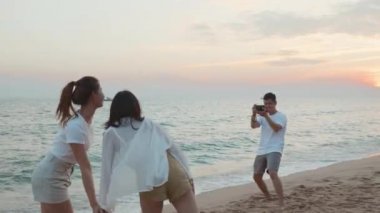 Mutlu arkadaşlar gülümsüyor ve poz veriyor, akıllı telefondan fotoğraf çekiyor ve tatilde tatilde gün batımında sahilde birlikte eğleniyorlar, genç Asyalı grup kadın ve erkek yaz gezisinde.