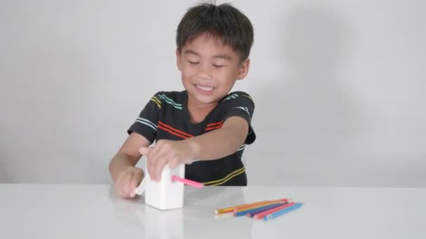 Kleine süße Junge 5-6 Jahre alt lächeln mit Bleistiftspitzer während der Hausaufgaben im Studio Schuss isoliert auf weißem Hintergrund, asiatische Kinder Vorschule schärfen Farbstifte, Bildungskonzept