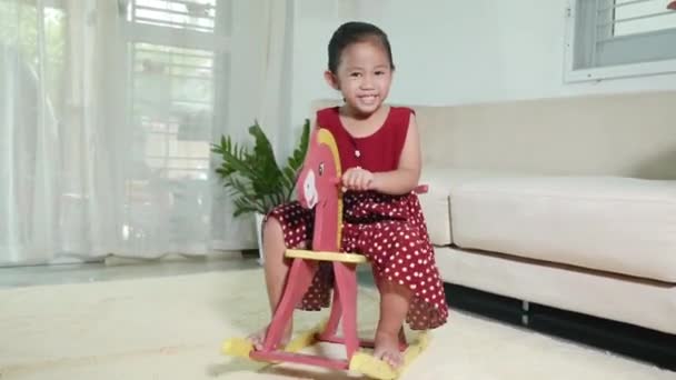 アジアの可愛い女の子スイングおもちゃの馬に乗って 笑顔の子供は リビングルームで自宅で馬のロッキングチェアを再生し 幸せな時間は 自己幼稚園就学前の間に再生する — ストック動画