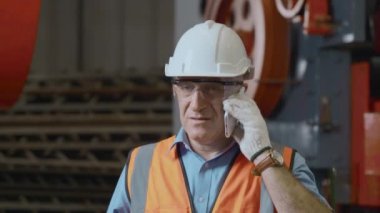 Kuasyalı teknisyen çalışırken güvenlik üniforması giyer. Metal levha fabrikası atölyesinde mühendislik ve konuşmak için cep telefonunu arar.