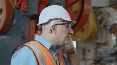 Kuasyalı teknisyen çalışırken güvenlik üniforması giyer. Metal levha fabrikası atölyesinde mühendislik ve konuşmak için cep telefonunu arar.