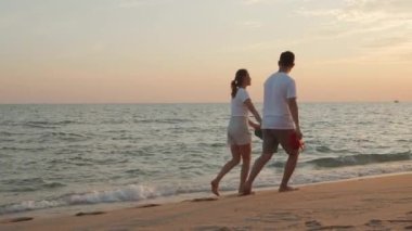 Tatil aşığı çift gün batımında kumsalda yürüyorlar, yaz tatilinde dinleniyorlar, aşık Asyalı erkek ve kadın kumlu sahil boyunca el ele yürüyorlar.