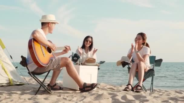 快乐的朋友在夏令营里弹吉他和拍掌玩得很开心 他们一起在沙滩度假时 在日落时分 野营帐篷附近的度假时间 年轻的亚洲女子和男子在户外旅行 — 图库视频影像