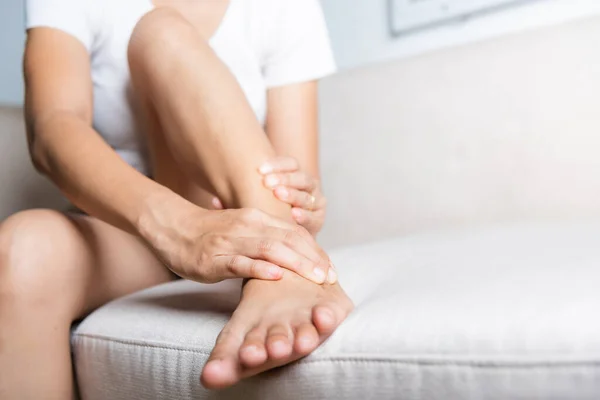 Foot pain, Asian woman sitting on sofa hold her ankle injury feeling pain in her foot at home, female suffering from feet ache use hand massage relax muscle from ankle interior, Healthcare and medical