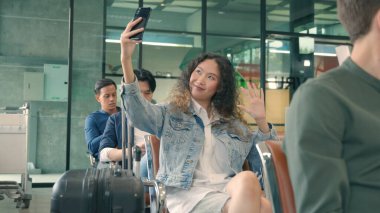 Seyahat tatilinin tadını çıkar. Mutlu Asyalı genç ve güzel kıvırcık saçlı kadın havaalanı terminalinde uçuşunu beklerken akıllı telefonuyla selfie çekiyor. Uluslararası havaalanında turist kız.