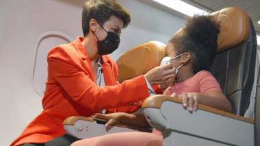 Anne, uçakta seyahat ederken yüz maskesi takan kıvırcık saçlı kıza koruma amaçlı yardım ediyor. Virüs salgını, yeni normal konsept, sağlık ve sağlık hizmetleri.