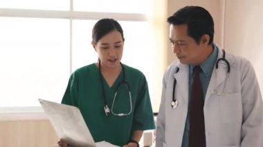 Hemşire ve kıdemli doktor hastane koridorunda pano tartışmalarıyla meşguller, doktor ve hemşire hastane koridorunda yürürken sağlık raporlarıyla konuşuyorlar.