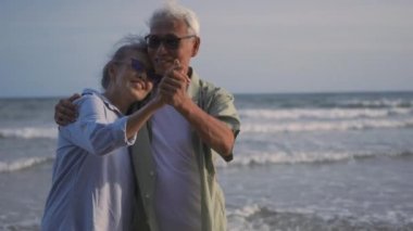 Mutlu Asyalı erkek ve kadın kıdemli çift gülümser dans eder plaj balayında dinlenir aile güneşli bir günde romantik yaşlılar yaz tatilinde seyahat eder emekliler için hayat sigortası yaptırır.