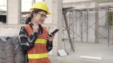 Ev inşaatında çalışırken planlama ve geliştirme projelerine takımla birlikte akıllı telefon uygulaması kullanarak koruyucu miğfer ve üniformalı Asyalı mimar mühendis kadın.