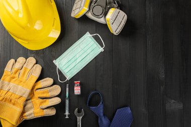 Patlak Coronavirus sırasındaki İşçi Bayramı 'nız kutlu olsun. Çeşitli inşaat malzemeleri elle kumanda donanımı, COVID-19 aşısı, siyah ahşap arka planda kopyalama alanı olan tıbbi yüz maskesi.