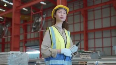 Güvenlik yeleği olan Asyalı kadın elinde tablet bilgisayarla fabrikadaki depolama deposuna doğru yürüyor profesyonel mühendis kontrol makinası çatıdaki metal levha depolama endüstrisinde yürüyor.