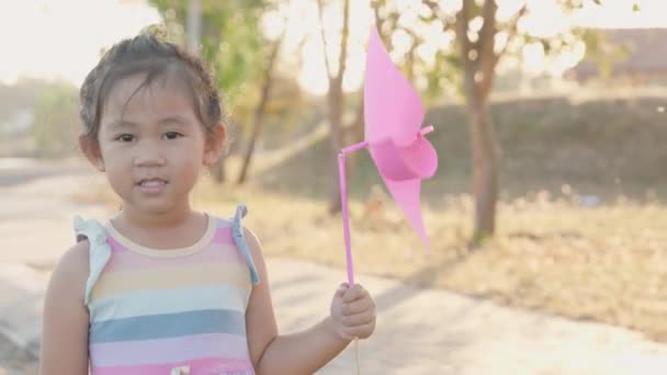 アジアの女の子遊びますとともにピンホイール 幸福小さな女の子笑顔で小さな風車や風車のおもちゃを手に夏の日に公園の庭で — ストック動画