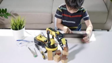 Asyalı küçük çocuk atölyede dijital tablet bilgisayar kullanıyor, çocuk ders programlama kontrol robotu kolu öğreniyor tahta bloğu kaldırıyor, teknoloji bilimi eğitimi alıyor.