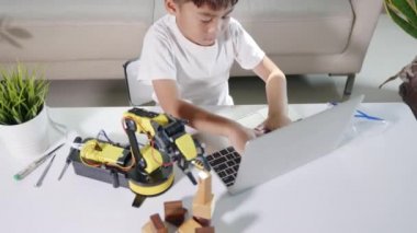 Asyalı küçük çocuk atölyede dizüstü bilgisayar kullanıyor. Robotik kol makinesini kontrol ediyor. Programlama kontrol robotu kolu için çocuk öğrenme programcısı.
