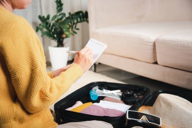 Making check list of things to pack for travel. Woman writing paper take note and packing suitcase to vacation writing paper list sitting on room, prepare clothes into luggage, Travel vacation travel