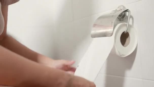手拉着卫生纸滚筒擦拭 女人坐在马桶上 她拿着并撕破了墙上的白色纸巾在浴室里擦拭 医疗保健理念 — 图库视频影像