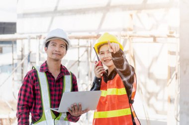 İnşaat alanında çalışan Asyalı mühendis erkek ve kadın ustabaşı dizüstü bilgisayar kullanıyor ve radyo, mühendislik bölümü bilgisayarları ve radyo ile konuşuyor.