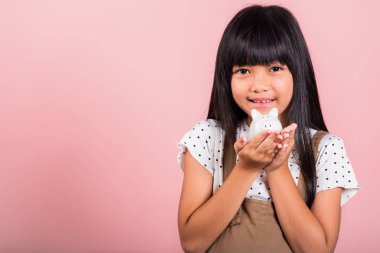 Asyalı küçük bir çocuk kumbarasını tutuyor ve pembe arka planda izole edilmiş stüdyo çekimlerine bakıyor, mutlu bir çocuk gibi gülümsüyor, tam bir domuz kumbarası, kişisel tasarruf