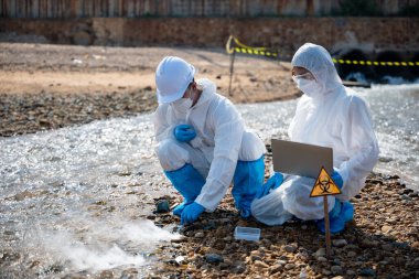Biyolog koruyucu kıyafet giyer ve maske endüstriden, sorunlu çevreden atık su numunesi toplar, ekolojist örneği incelemek ve veri kaydetmek için ölü balığı laptopa, toksik suya götürür.