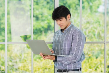 Asyalı işadamı gülümsüyor ve ofisin penceresinin yanındaki dizüstü bilgisayarda çalışıyor. Ofis lobisinde bilgisayarla buluşan yakışıklı bir adamın portresi.