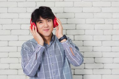Asyalı yakışıklı bir gencin portresi. Tuğla duvar arkasında kulaklıkla müzik dinlerken gülümsüyor. Mutlu genç adam gülümsüyor. Kırmızı kulaklıklı mutlu adam.