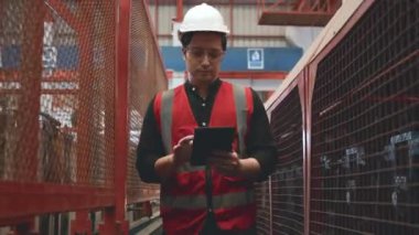 Güvenlik yeleği olan Asyalı işçi elinde tablet bilgisayarla fabrikanın bakımı için kontrol makinesine gidiyor, profesyonel mühendis kontrol mekanizmaları kontrol ediyor metal levha depolama endüstriyel yürüme