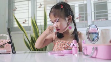 Makyaj yapan Asyalı komik küçük kız akıllı telefona bakıyor ve maskara kullanıyor, kadın olmak için öğrenme aktivitesi, sosyal medya blogcusu kozmetik ile video kaydı, online özel ders.