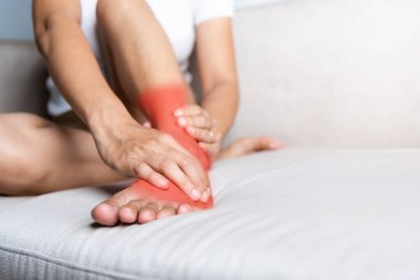 Ayak ağrısı, kanepede oturan Asyalı bir kadın ayak bileği incinmiş, ayaklarında ağrı var, ayak masajı yaptırıyor, el masajı yaptırıyor, ayak bileğinin iç kısmından kas gevşetiyor, sağlık ve sağlık sigortası yaptırıyor.
