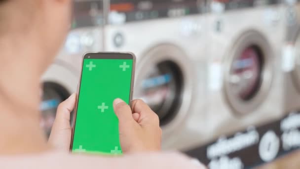 洗濯機作業中に洗濯機でデジタルスマートフォンを使用する女性 セルフサービスの洗濯設備 スマートフォンの緑の画面で洗濯するための女性の設定アプリケーション — ストック動画