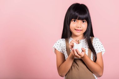 Asyalı küçük bir çocuk kumbarasını tutuyor ve pembe arka planda izole edilmiş stüdyo çekimlerine bakıyor, mutlu bir çocuk gibi gülümsüyor, tam bir domuz kumbarası, kişisel tasarruf