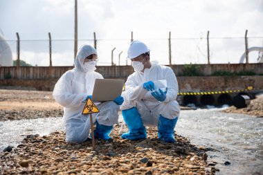 Ekolojist ölü balığı incelemek ve dizüstü bilgisayara veri kaydetmek için örnek alıyor, biyolog koruyucu giysi giyiyor ve endüstriden, sorun ortamından gelen atık su örneklerini maskeliyor