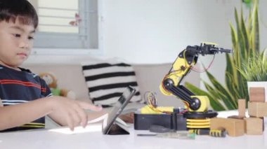 Asyalı küçük çocuk atölyede dijital tablet bilgisayar kullanıyor, çocuk ders programlama kontrol robotu kolu öğreniyor tahta bloğu kaldırıyor, teknoloji bilimi eğitimi alıyor.