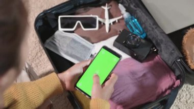 Akıllı telefonlu boş yeşil ekran kullanan ve hafta sonu tatili için bavul hazırlayan kadın, havaalanına gitmeden önce cep telefonuyla turizm sitesindeki kadın kontrol bilgileri