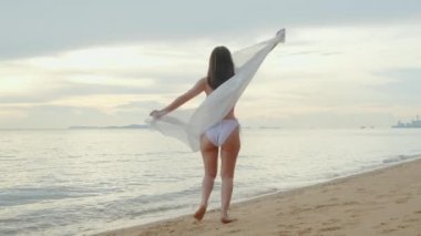 Genç, güzel bir kadın bikini mayo giyip sarong üfleyerek dans ediyor ve tropikal plajda vücut değiştiriyor mutlu Asyalı kadınlar dışarıda tatilde eğleniyor yaz tatili tatillerinde dinleniyor.