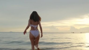 Geriye bakış, genç güzel bir kadın bikini giyiyor tropikal plaja koşuyor ve zıplıyor elini kaldırıyor mutlu Asyalı kadın eğleniyor ve tatilde gün batımının dışında zıplıyor yaz tatili
