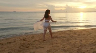 Güzel, genç bir kadın rüzgarda bikini mayo giyer, eşarp takar, tropikal plajlarda yürür ve altın gün batımında özgürlük için elini kaldırır.