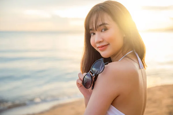 Portrait of young woman in white bikini swimsuit standing posing and holding sunglasses on tropical beach, Happy beautiful Asian girl in swimwear on summer holiday travel