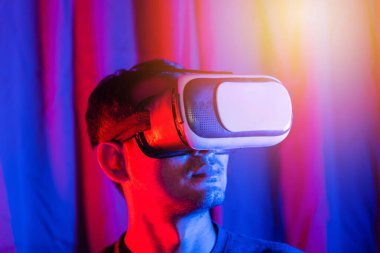 Asyalı yakışıklı bir adam VR kaskı takıyor. Evde duygusal, mor ve mavi arka planda sanal gerçeklik gözlüğü takıyor. Gerçekliği tecrübe ediyor.
